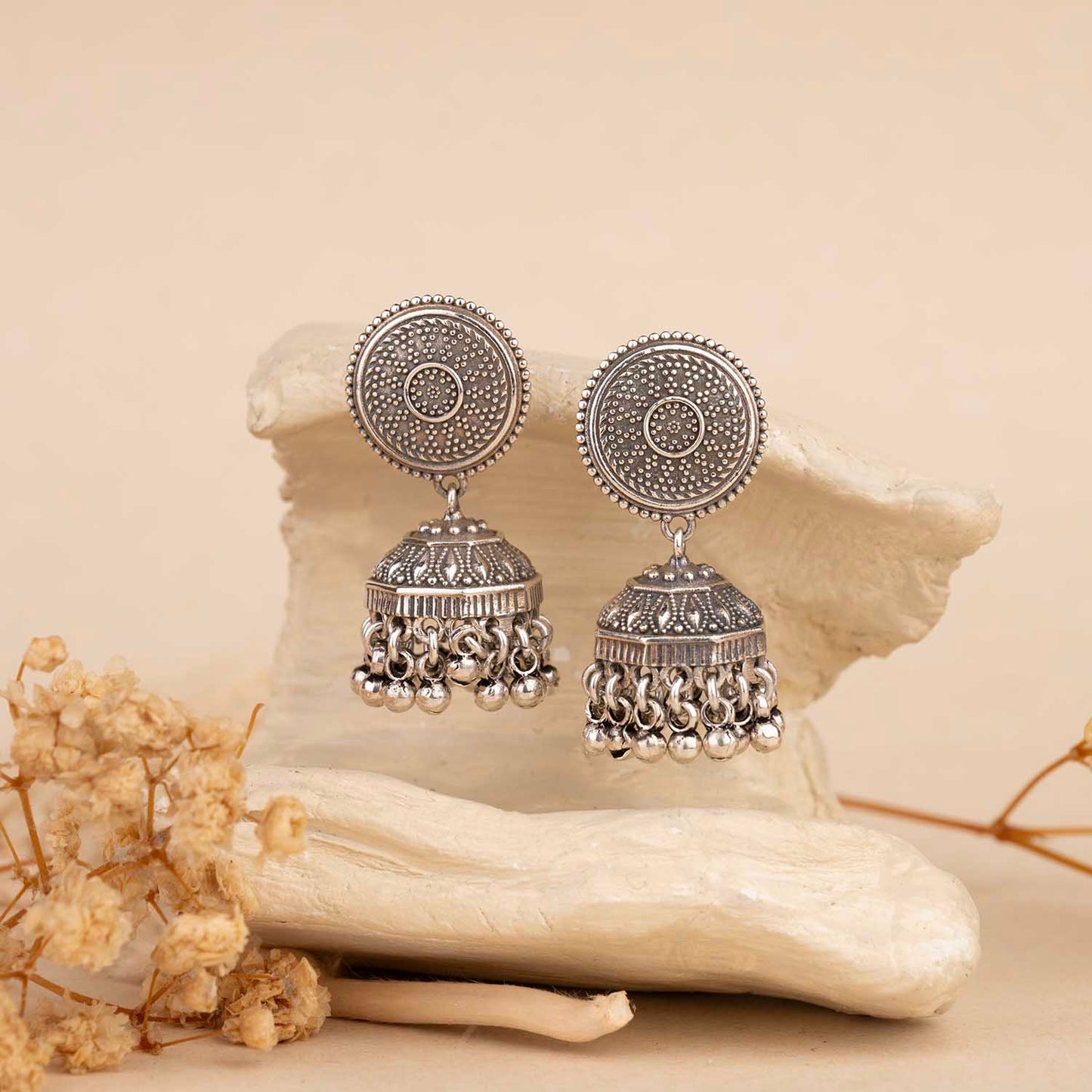 Pair of silver earrings on a textured beige surface with dried flowers.
Silver Jhumka