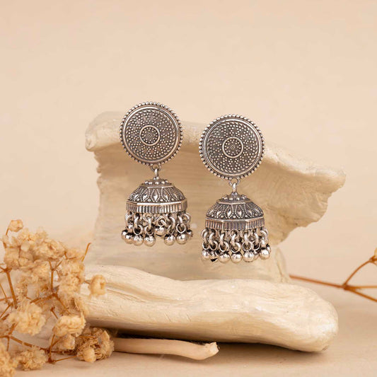 Pair of silver earrings on a textured beige surface with dried flowers.
Silver Jhumka
