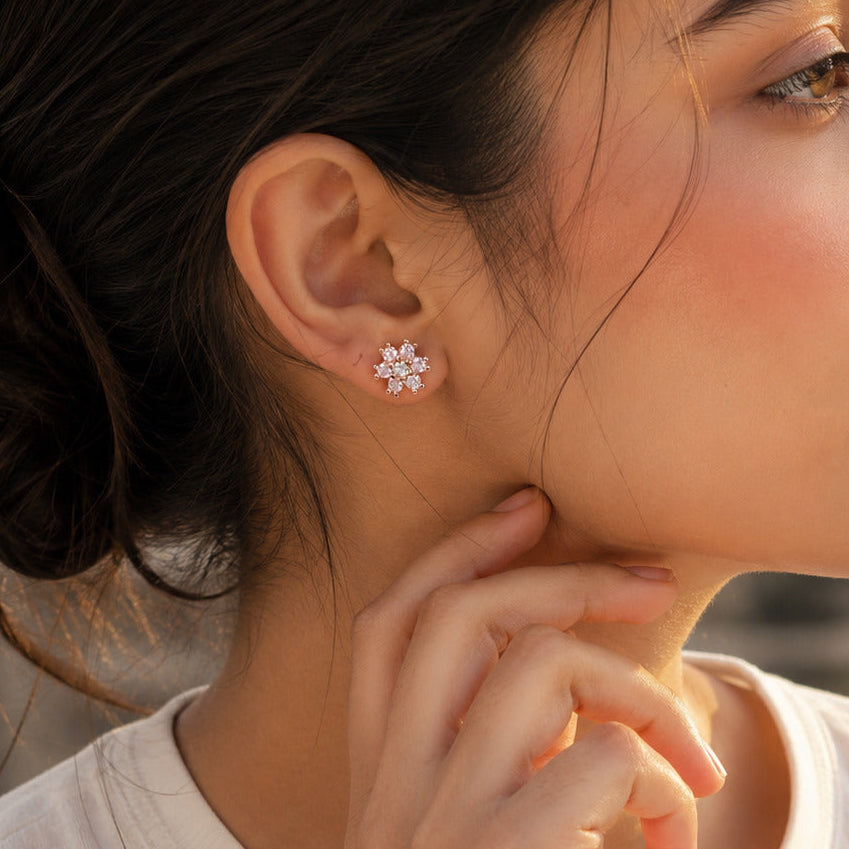 A close-up profile shot of a woman wearing a small, sparkling snowflake-shaped stud earring. She has dark hair tied back in a bun and is wearing a white t-shirt, with warm sunlight highlighting her face and ear.